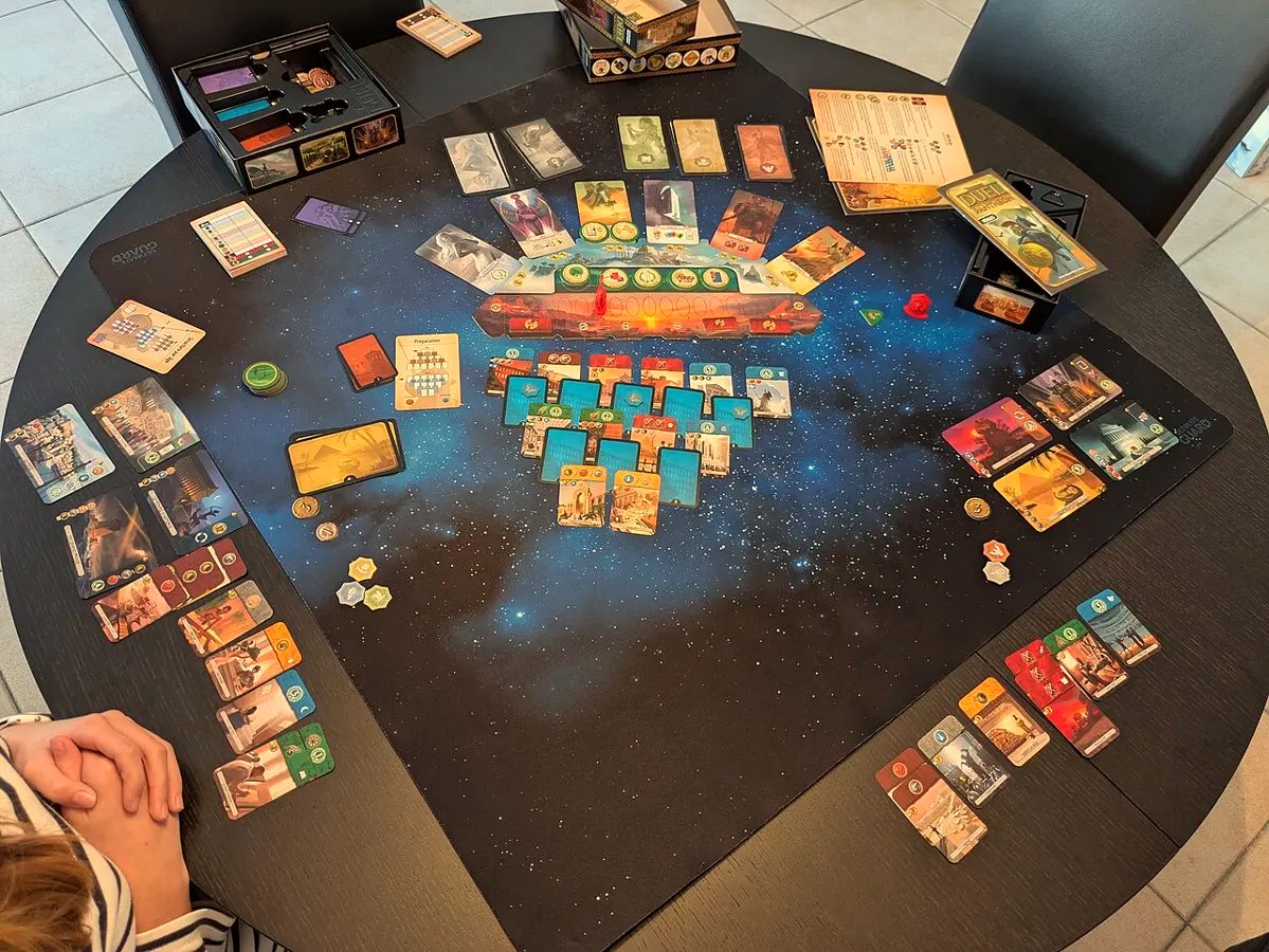 A game of 7 Wonders Duel laid out on a round table, cards in rows, a player's hands resting at the edge