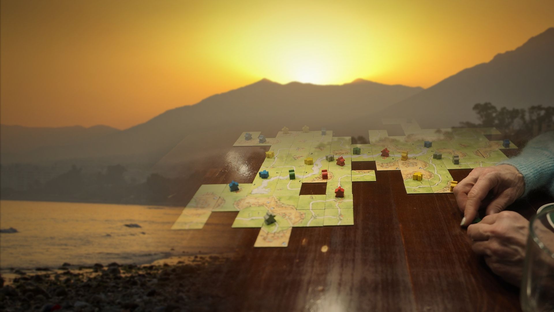 Illustrative composite: a game of Carcassonne in play, hands at the table, over a sunset on the Marbella coast