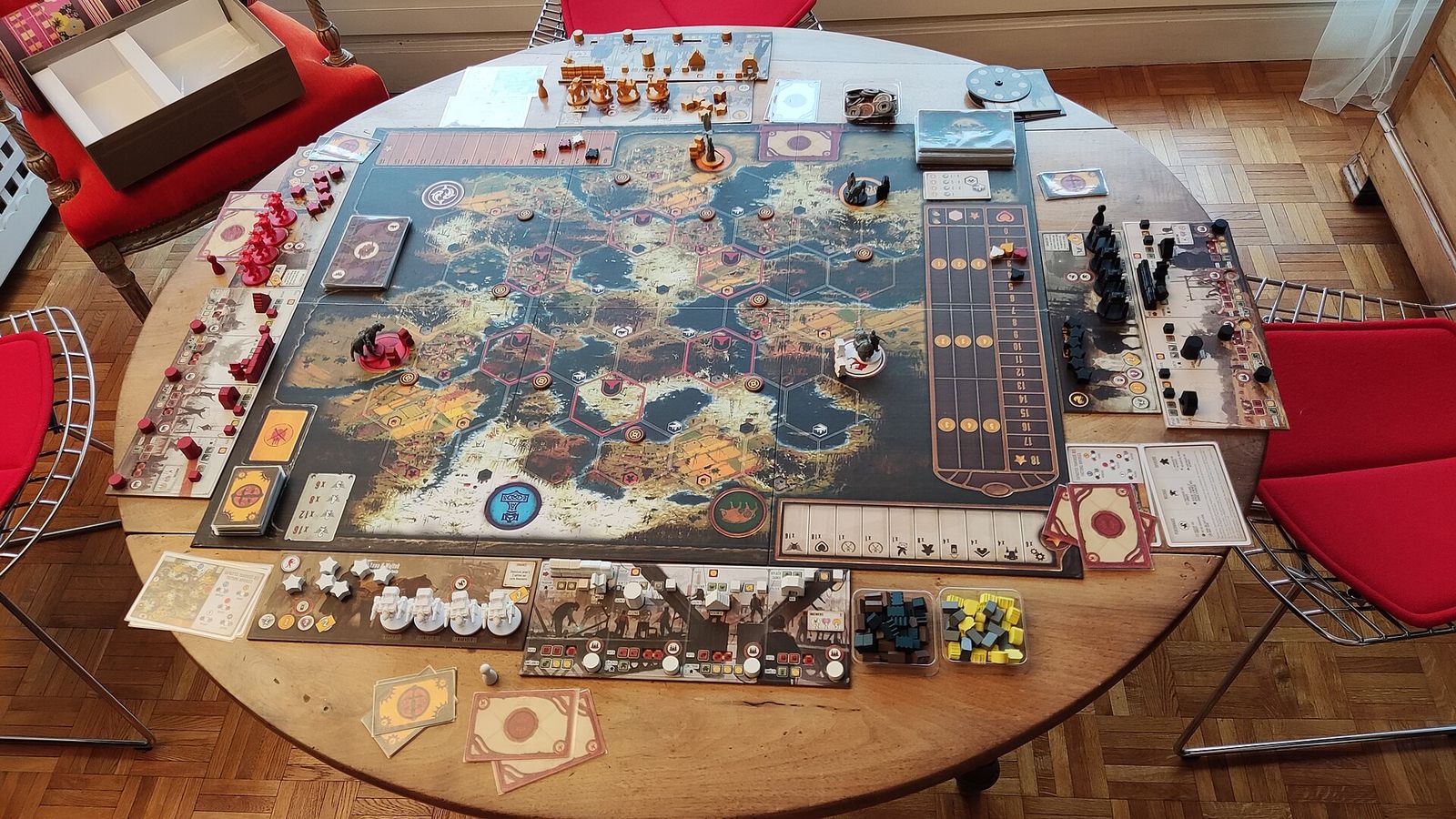 A game of Scythe set up on a round wooden table: the board, cards, miniatures and tokens in place, red chairs pulled up around it